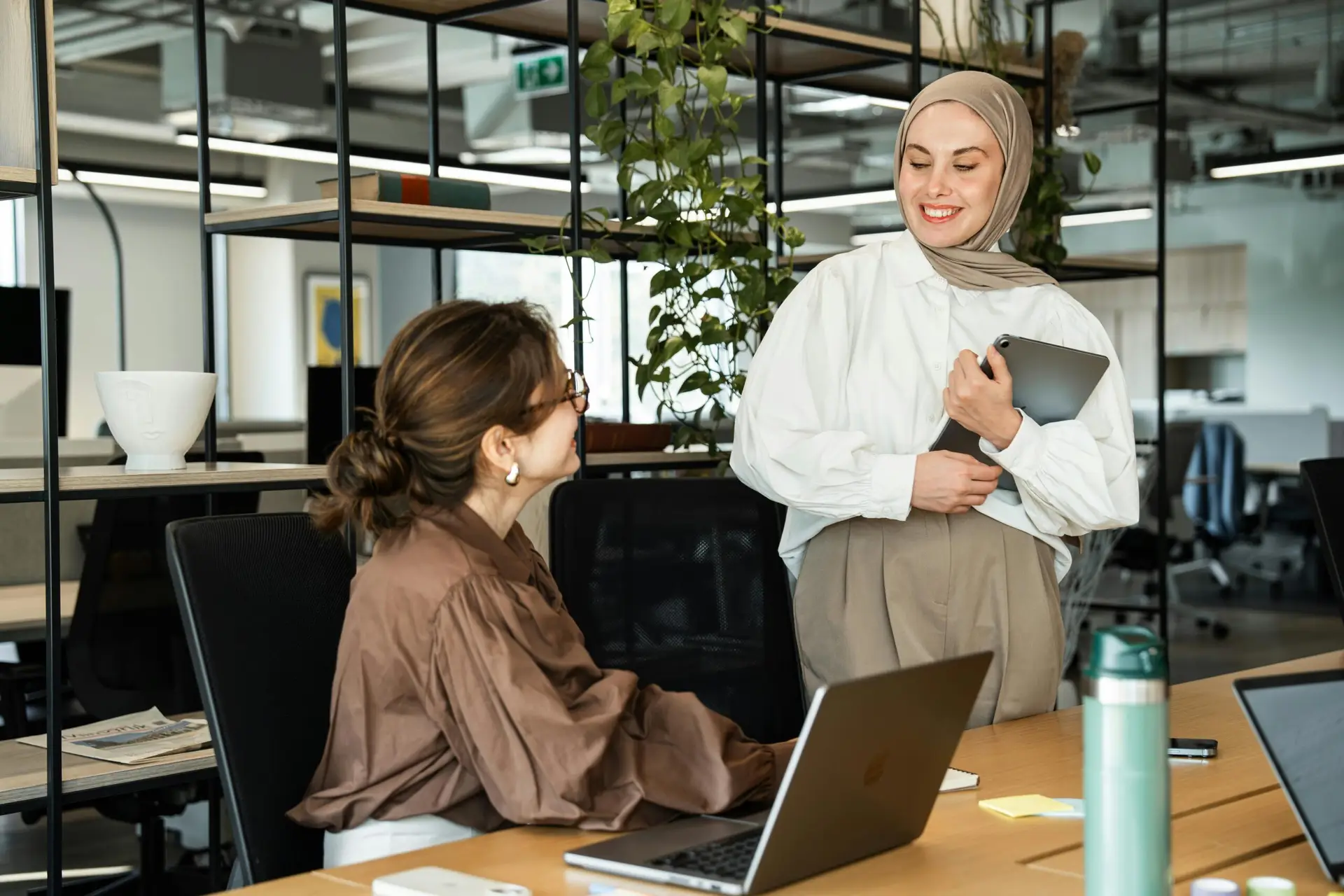 Two professional women in a modern office setting engaging in a positive interaction using laptops.